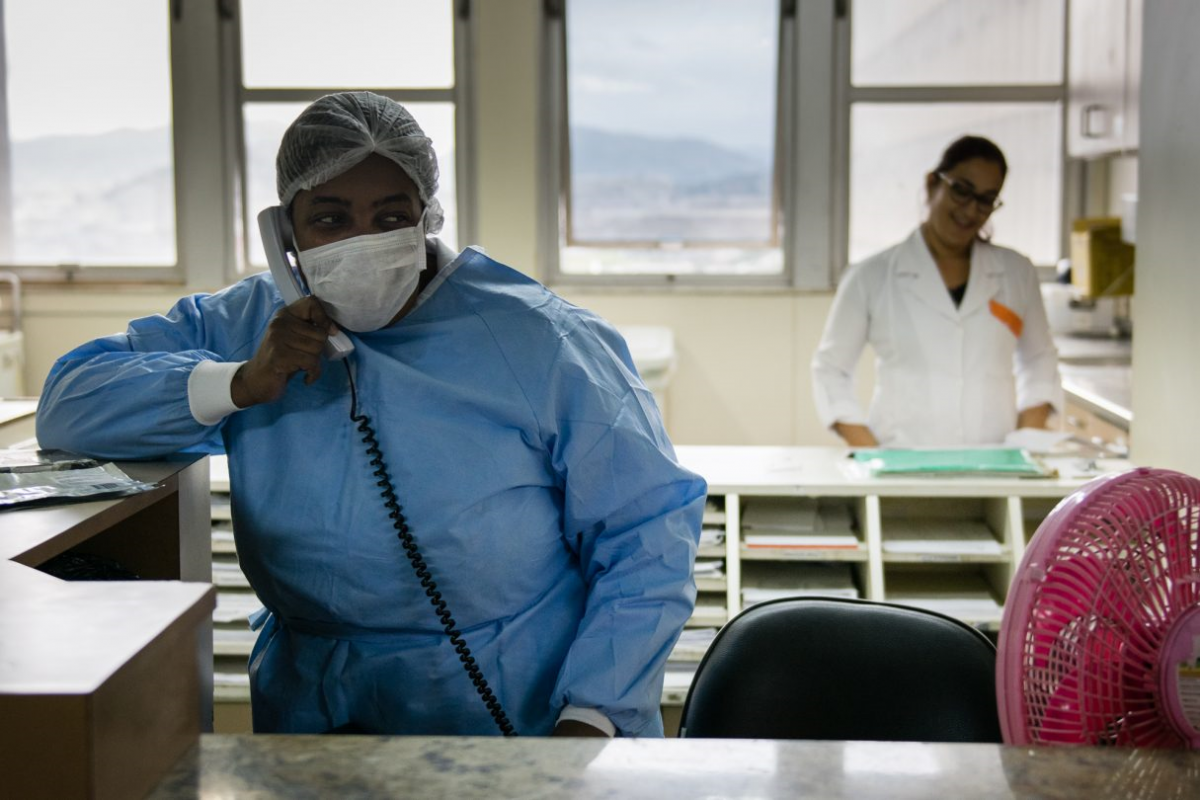 Uma enfermeira em primeiro plano usa uniforme e máscara enquanto atende o telefone.Em segundo plano, outra mulher trabalha.