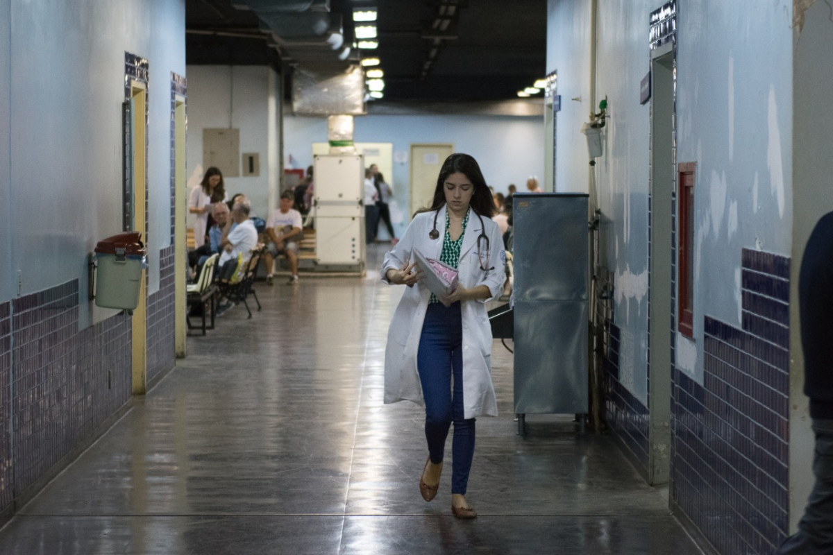 Mulher de jaleco caminha no corredor do Hospital Universitário. No fundo da imagem, pacientes aguardam sentados em cadeiras.