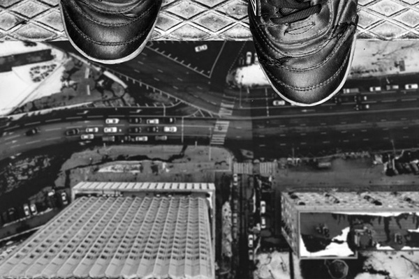 Imagem em preto e branco da ponta de dois pés calçados em cima de um prédio e com a visão da rua lá embaixo, simbolizando um abismo ou suicídio.