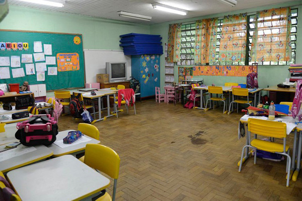 Imagem colorida. Sala de aula com mesas e cadeiras. Quadro verde ao fundo com exposição de trabalhos escolares e janela a direita.