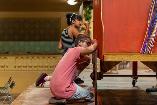 Sobre um palco, duas pessoas pintam uma estrutura de madeira que servirá como cenário de uma ópera