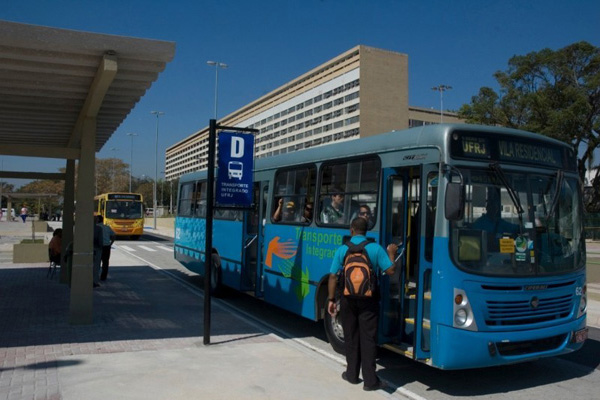 Em uma parada de ônibus localizada em frente ao prédio da Reitoria, pessoas entram em um ônibus azul e outras aguardam o carro seguinte