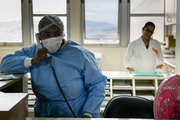 Imagem ilustrativa da matéria. Em primeiro plano, mulher negra com vestimenta de enfermeira está em um hospital e atende ao telefone. Ao fundo, desfocada, outra mulher com jaleco branco sorri enquanto trabalha