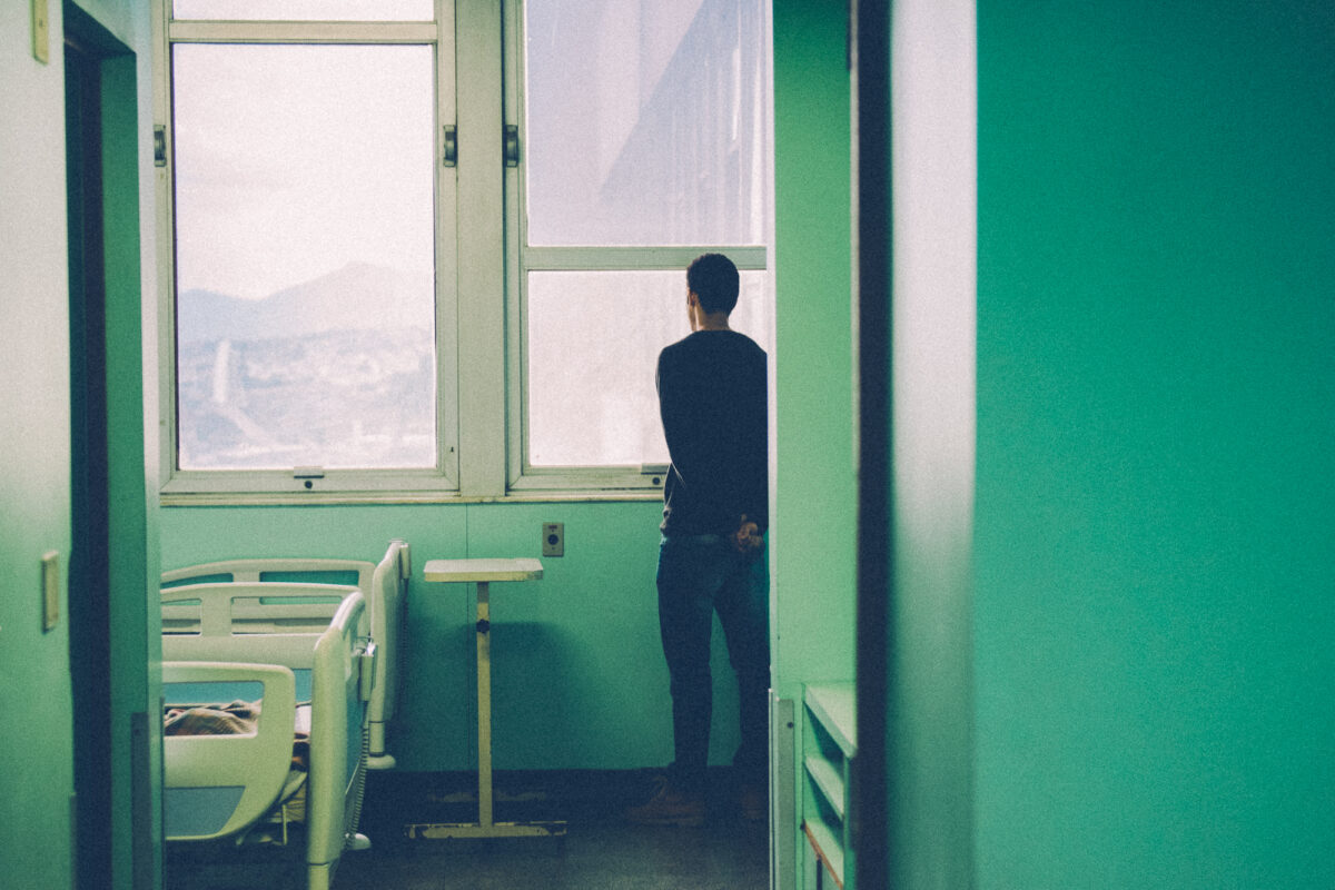 Homem de costas olha pela janela de quarto no Hospital Universitário