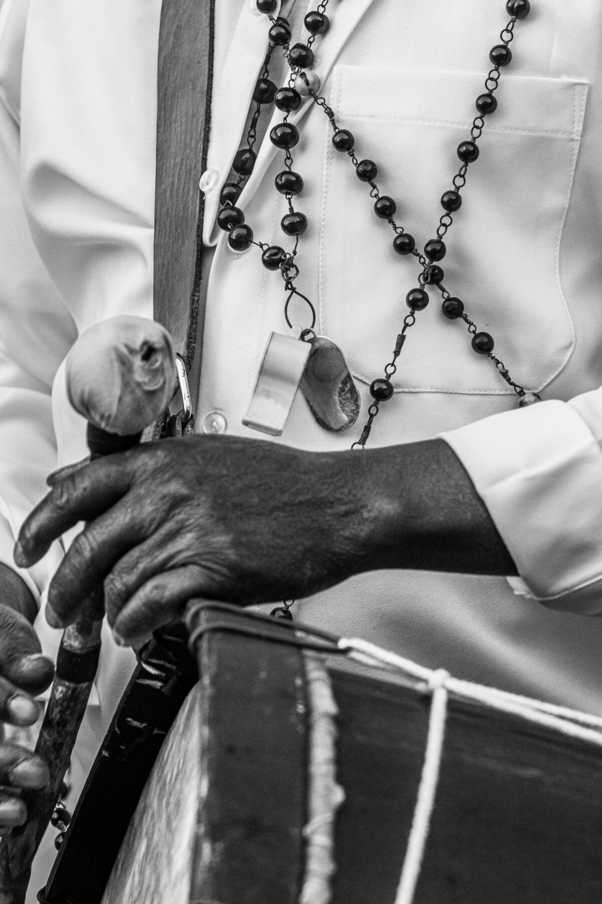 Mãos de um homem negro idoso tocam um tambor. Ele usa roupa branca e colares de conta.