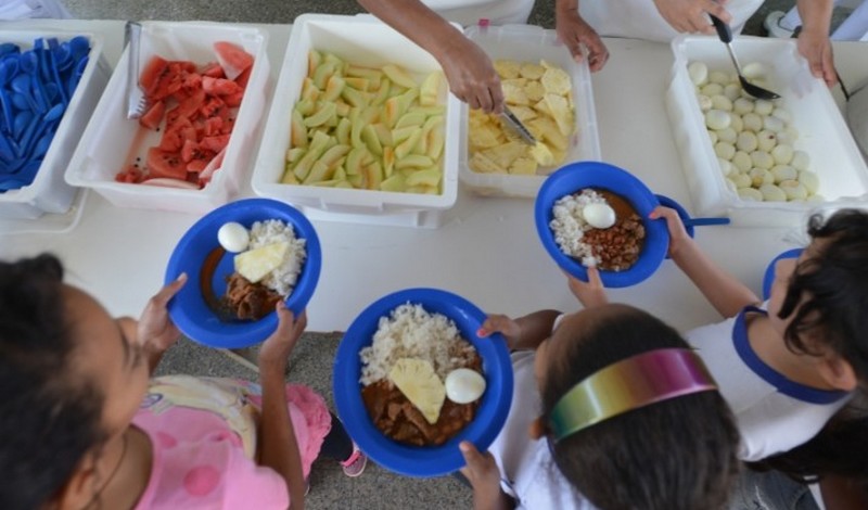 Crianças recebem a merenda na escola. O destaque da foto está na comida: arroz, feijão, ovos, melão, melancia e abacaxi
