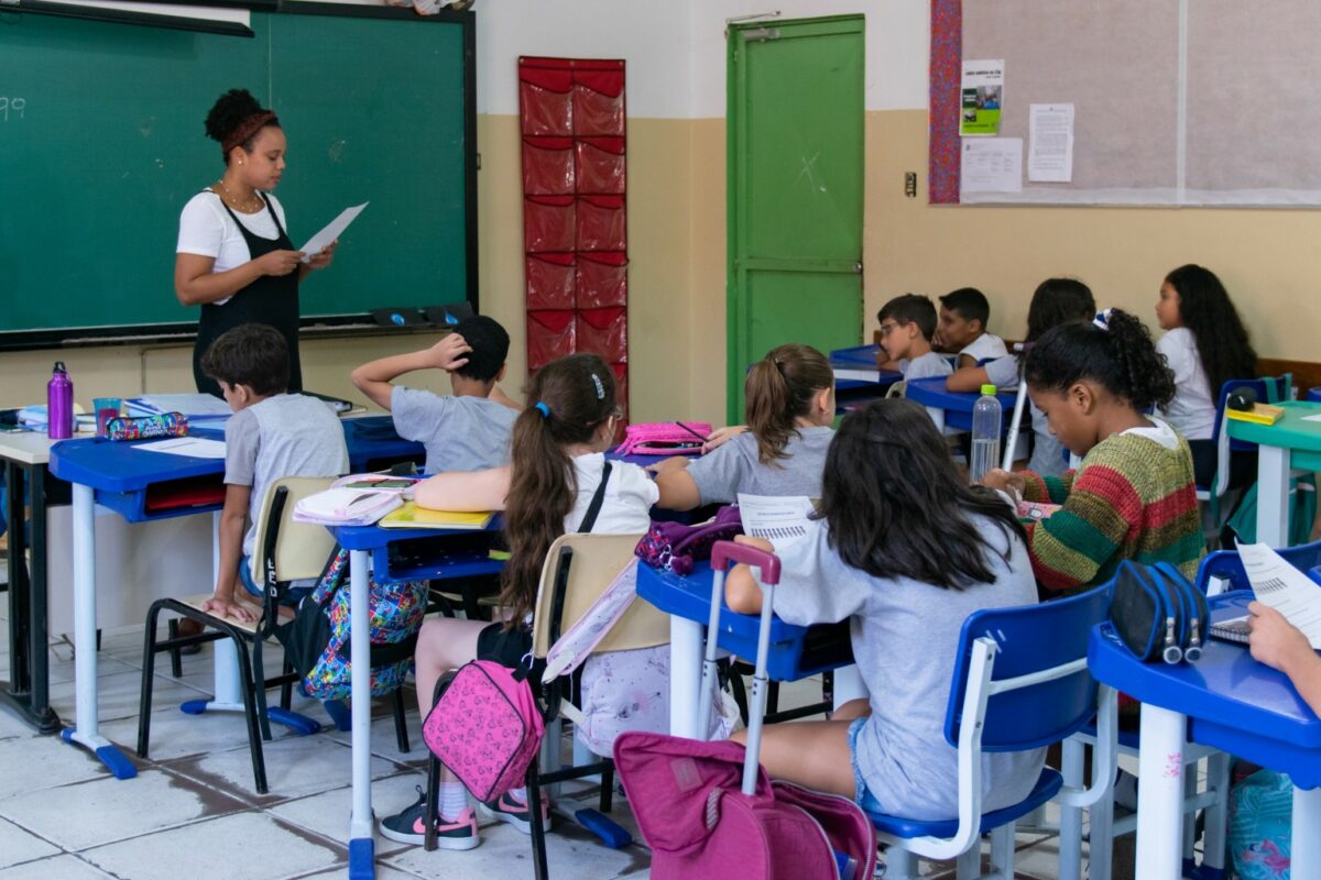 Crianças sentadas em sala de aula, com professora ministrando aula de pé