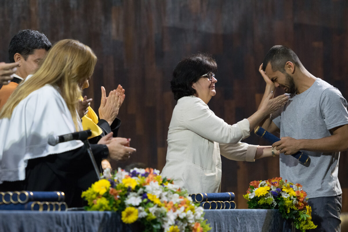 Em uma cerimônia, um estudante recebe seu diploma e se emociona. Uma professora o acolhe