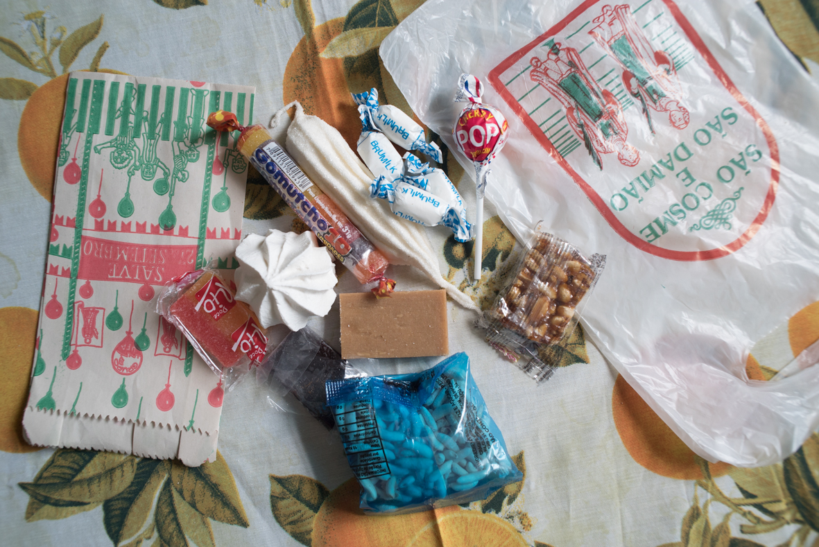 Fotografia em primeiro plano de um apanhado de doces e saquinhos distribuídos na festa de Cosme e Damião. Entre os doces há suspiro, maria-mole, jujuba, balinha de coco, pirulito e pipoca doce