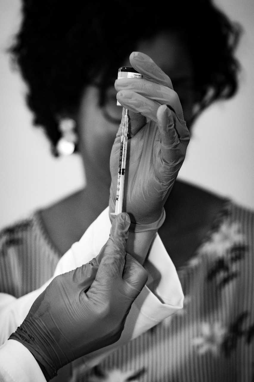 Em preto e branco, aparece em destaque a mão de uma pessoa que veste um jaleco e segura uma seringa de injeção, Ao fundo, aparece uma mulher negra