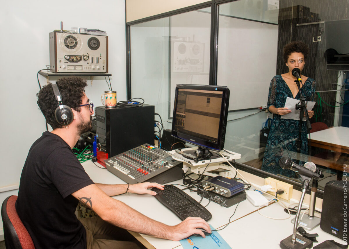 Em um estúdio de gravação, um homem opera equipamentos e, em frente a ele, separada por um vidro, uma mulher fala ao microfone
