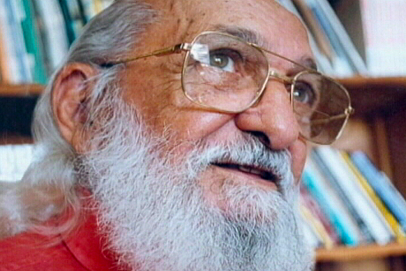 Imagem fechada no rosto de Paulo Freire, que olha para o alto. Ao fundo, uma estante de livros.