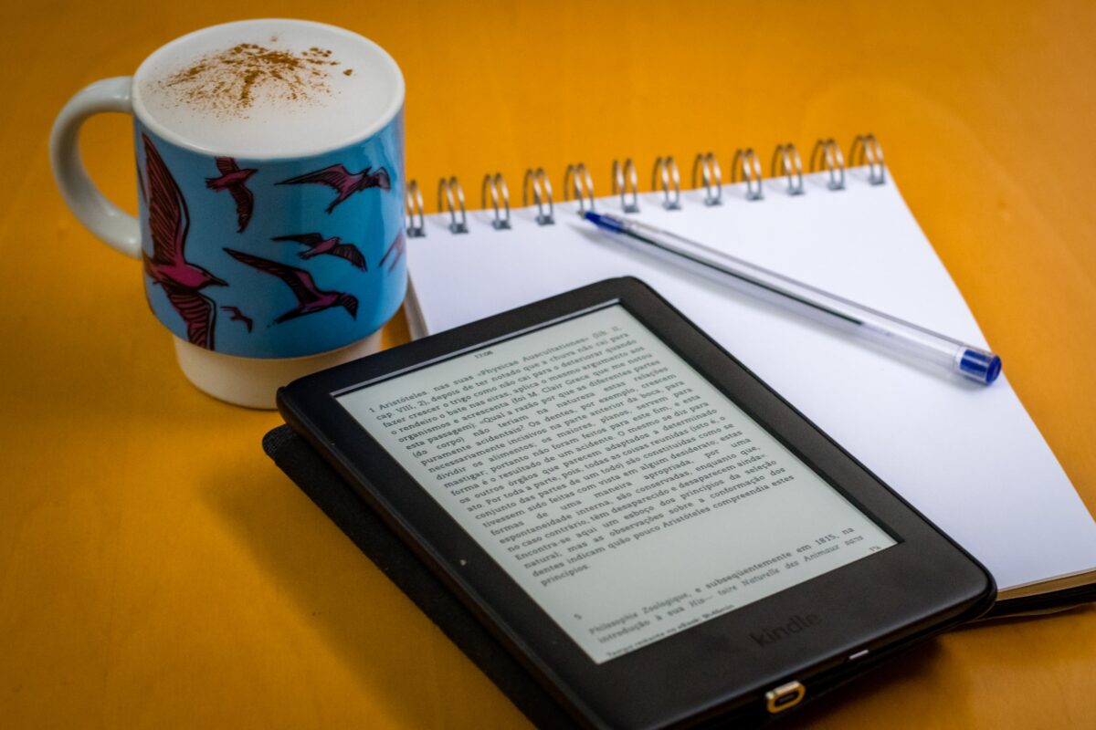 Sobre uma mesa amarela, uma caneca azul com café, um bloco de notas com uma caneta e um leitor digital com um e-book