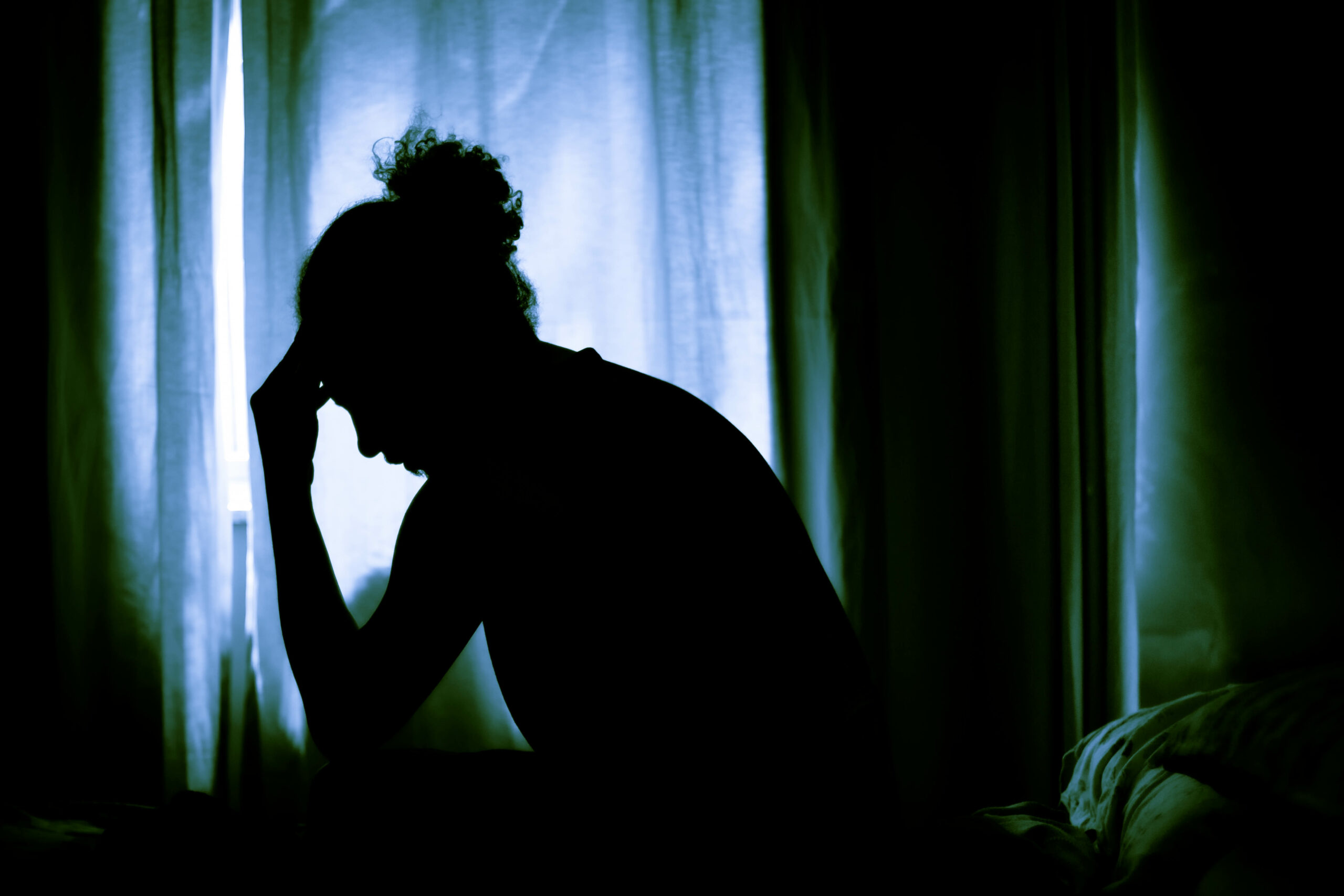 Em um ambiente escurecido pelas cortinas, uma pessoa está sentada diante de uma janela, cabisbaixa, com uma das mãos aparando a testa
