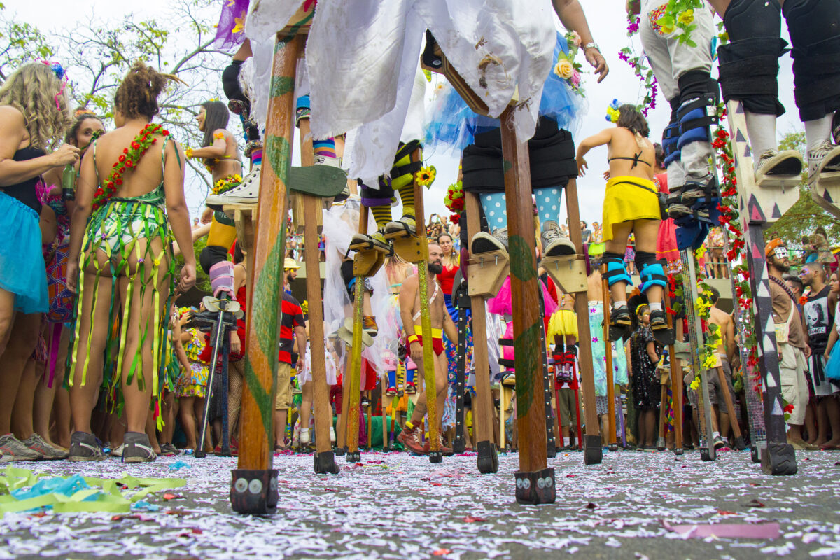 Foliões em um bloco de rua de carnaval. Em primeiro plano, pessoas usando pernas de pau. Em segundo pessoas fantasiadas.