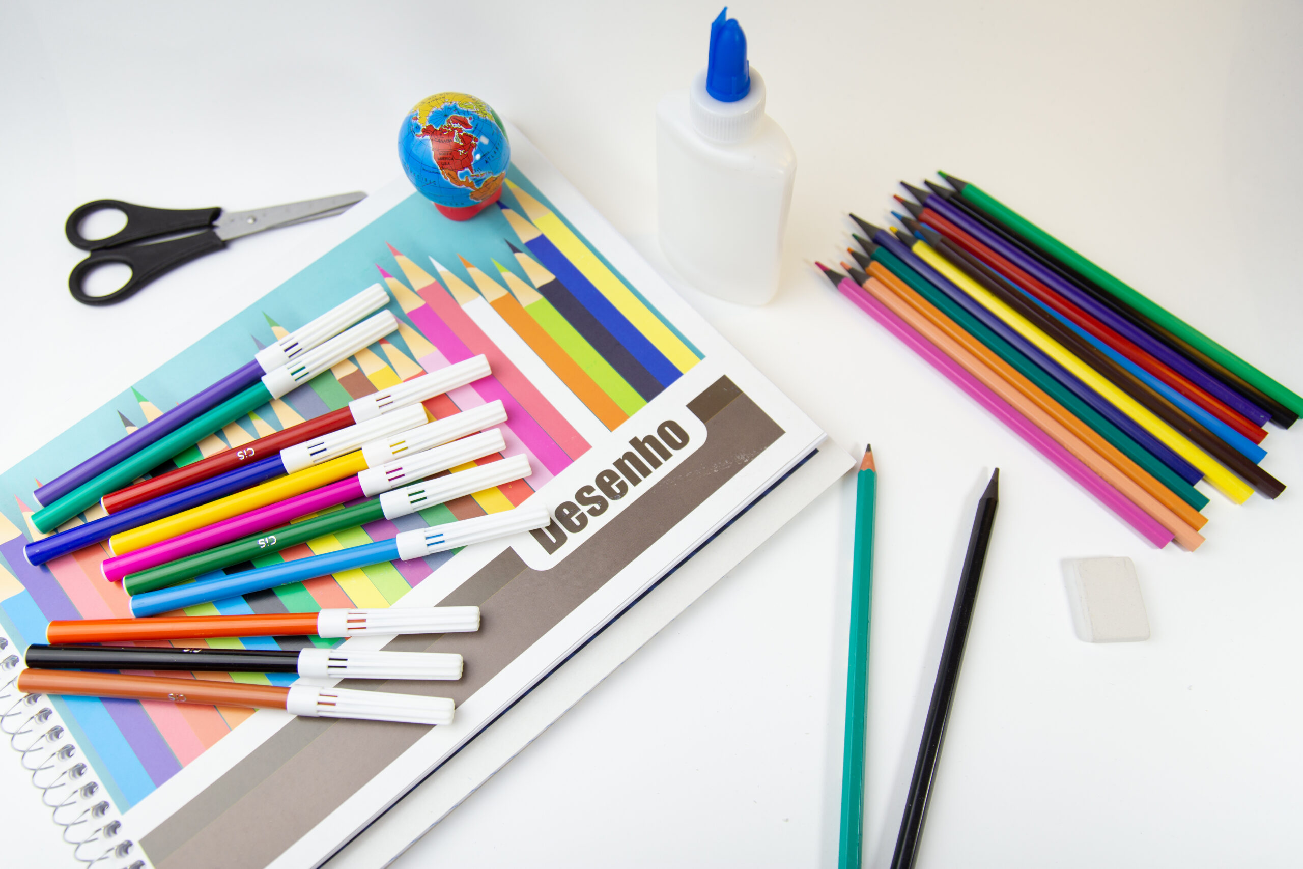 Sobre uma mesa branca, há canetinhas, lápis de cor, tesoura, cola e um caderno escrito desenho.