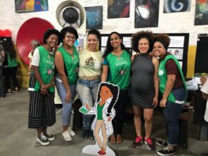 Seis mulheres negras posam para foto junto à mascote do projeto, uma menina negra cientista.