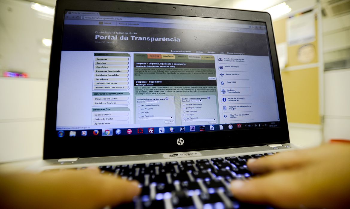Mãos humanas manuseiam um teclado de computador e, ao fundo, uma tela indica a página da internet que está sendo visitada: o Portal da Transparência do Governo Federal
