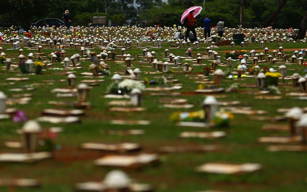 Em um cemitério, dezenas de lápides, decoradas com flores, se alinham no gramado. Ao fundo, há¡ duas pessoas agachadas, duas em pé e uma mulher caminha carregando um guarda-chuva