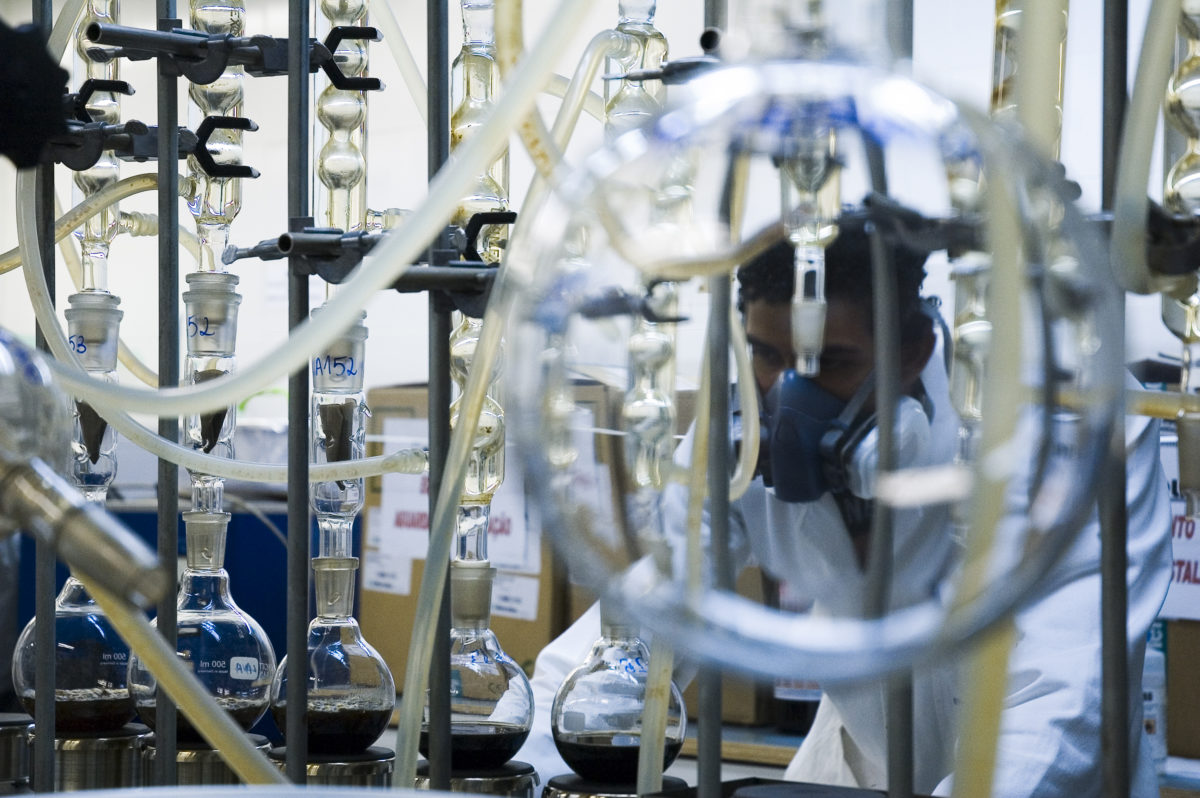 Em um laboratório de quí­mica, ao fundo, há um pesquisador de jaleco. Sua imagem se mistura à dos balões de fundo redondo distribuí­dos ao longo de uma bancada.