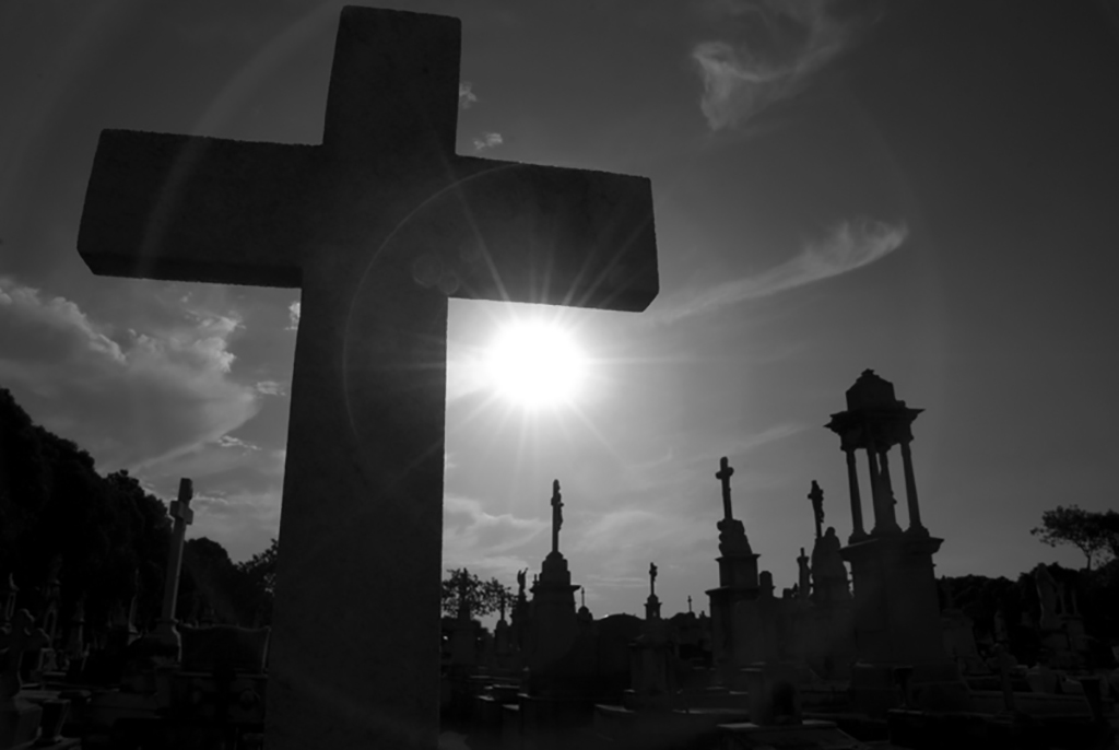 Foto em preto e branco mostra, em primeiro plano uma cruz. Ao fundo, túmulos no cemitério do Caju