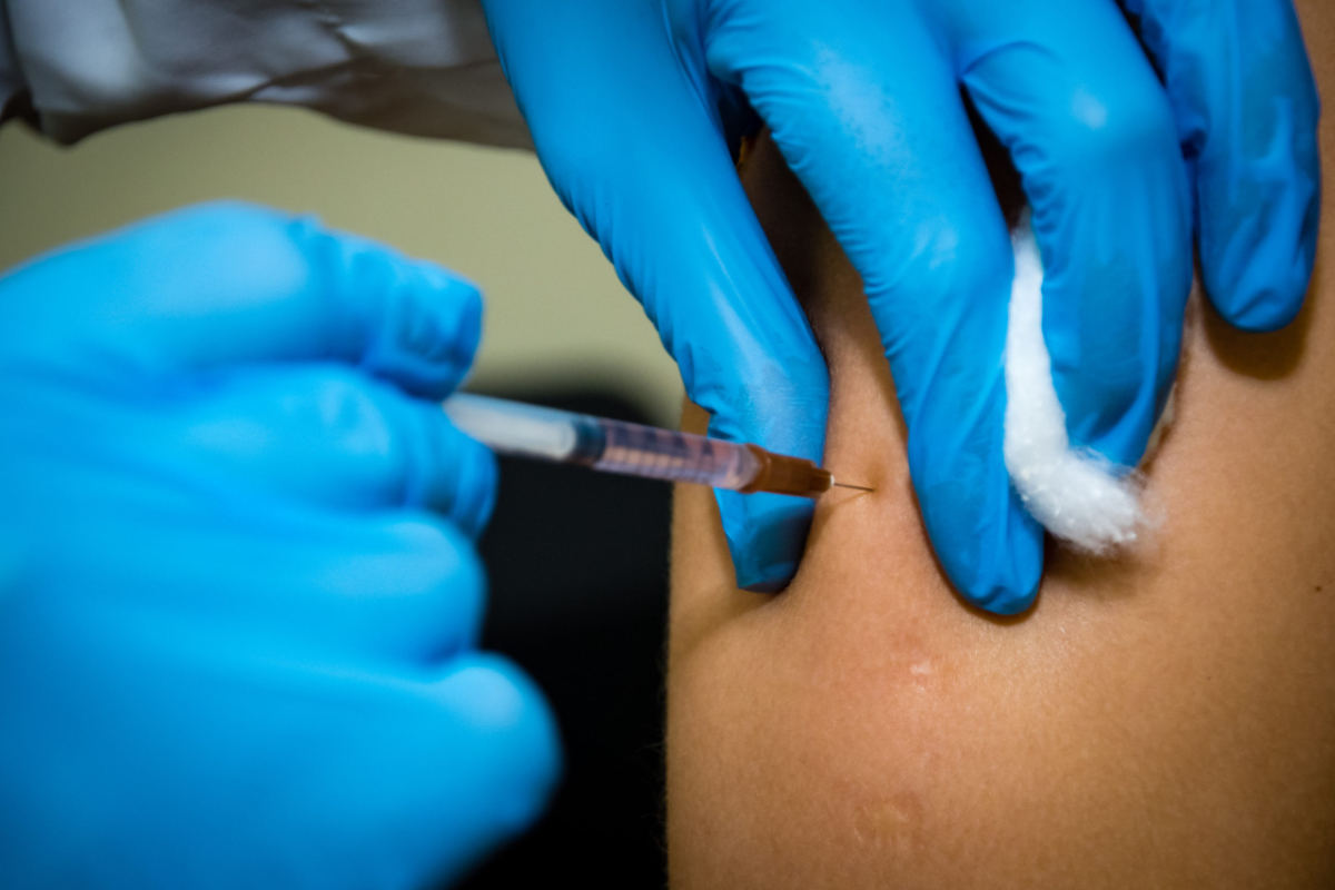 Mãos com luvas azuis aplicam vacina no braço de uma pessoa