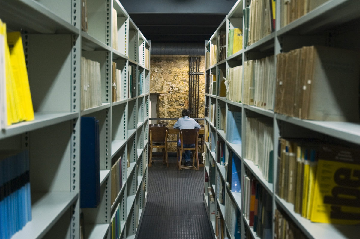 Corredor de uma biblioteca em meio a estantes com livros e documentos. Ao final do corredor, um estudante está sentado à mesa, de costas.