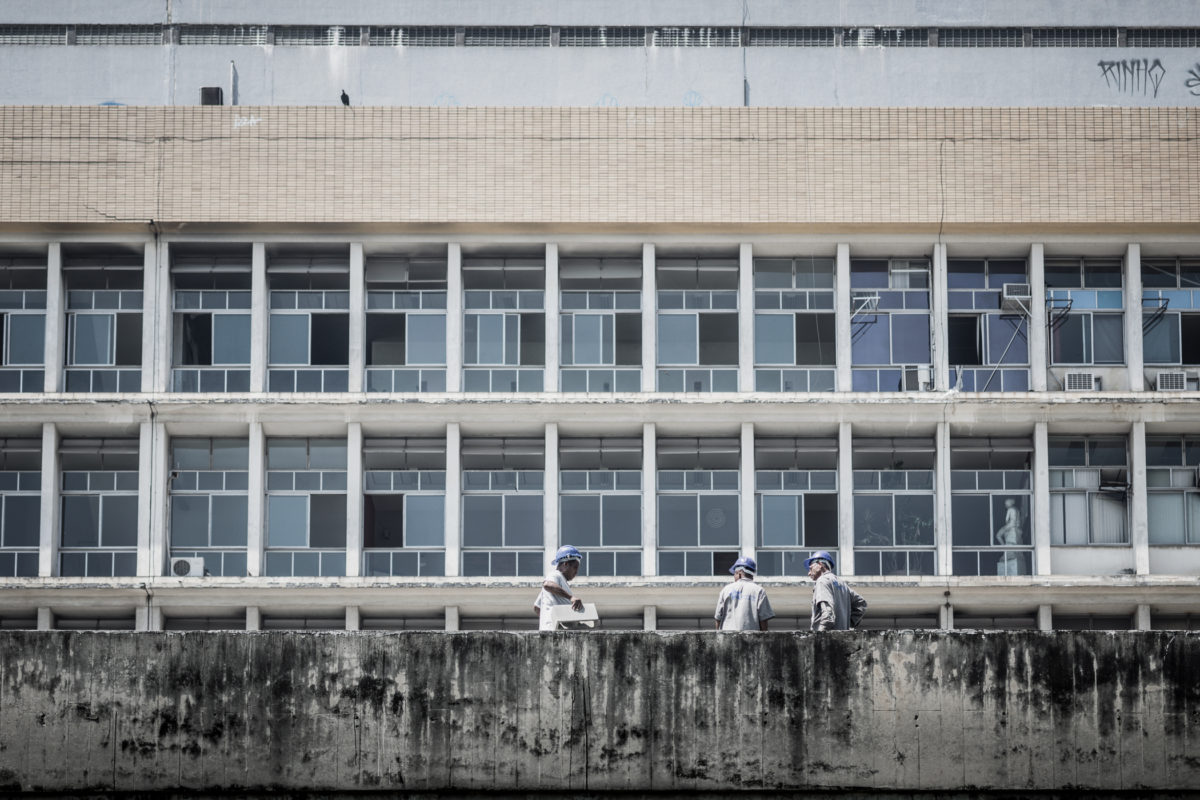 Três homens trabalham no terraço de um edifício. Ao fundo, um prédio mais alto, com várias vidraças quebradas, se impõe