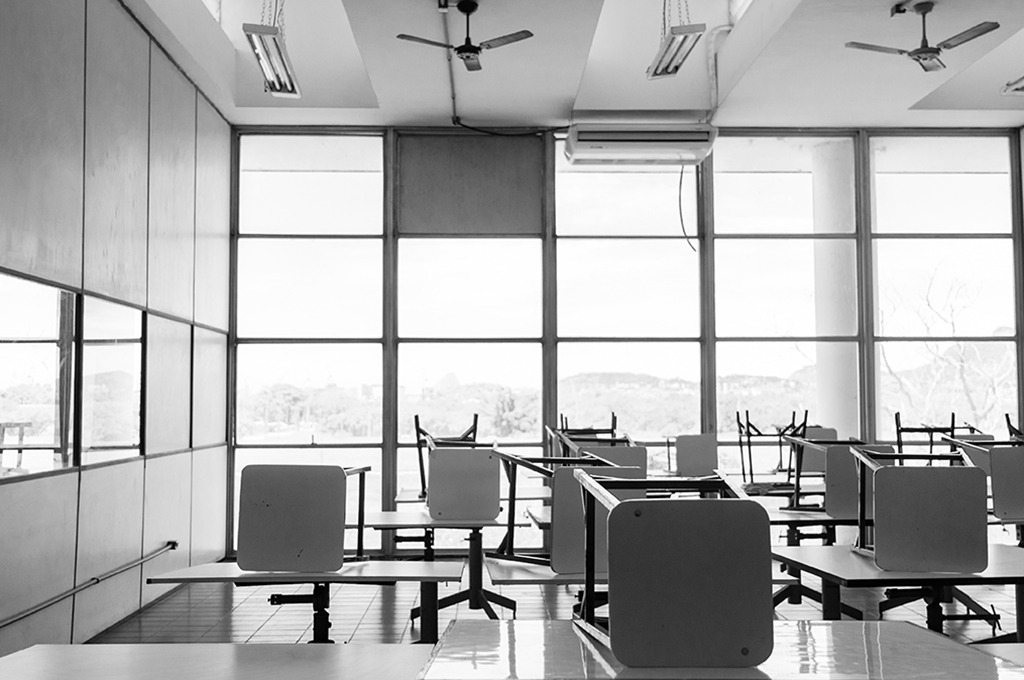 Fotografia em preto e branco de uma sala de aula vazia. As cadeiras estão sobre as mesas.