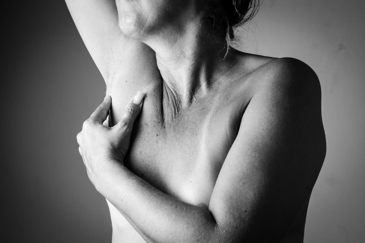 Foto em preto e branco mostra o dorso de uma mulher nua. Seu braço direito está levantado, enquanto a mão esquerda apalpa a axila, em um gesto de autoexame.