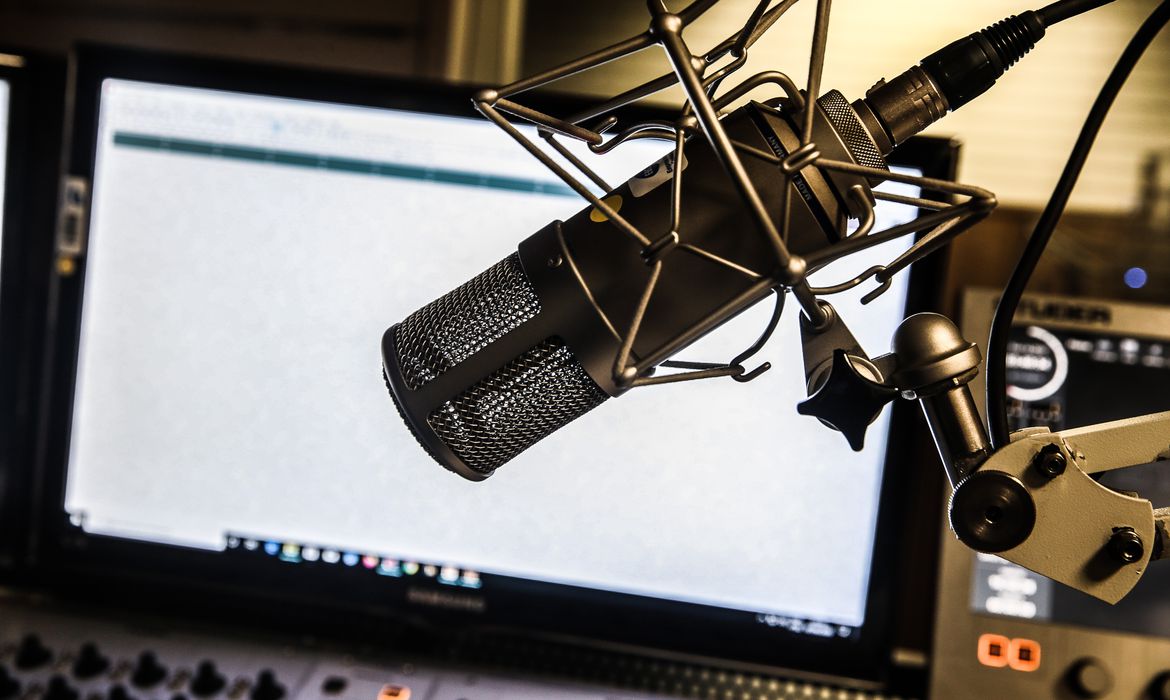 Fotografia de um estúdio de rádio: em primeiro plano, um microfone; ao fundo, um computador.