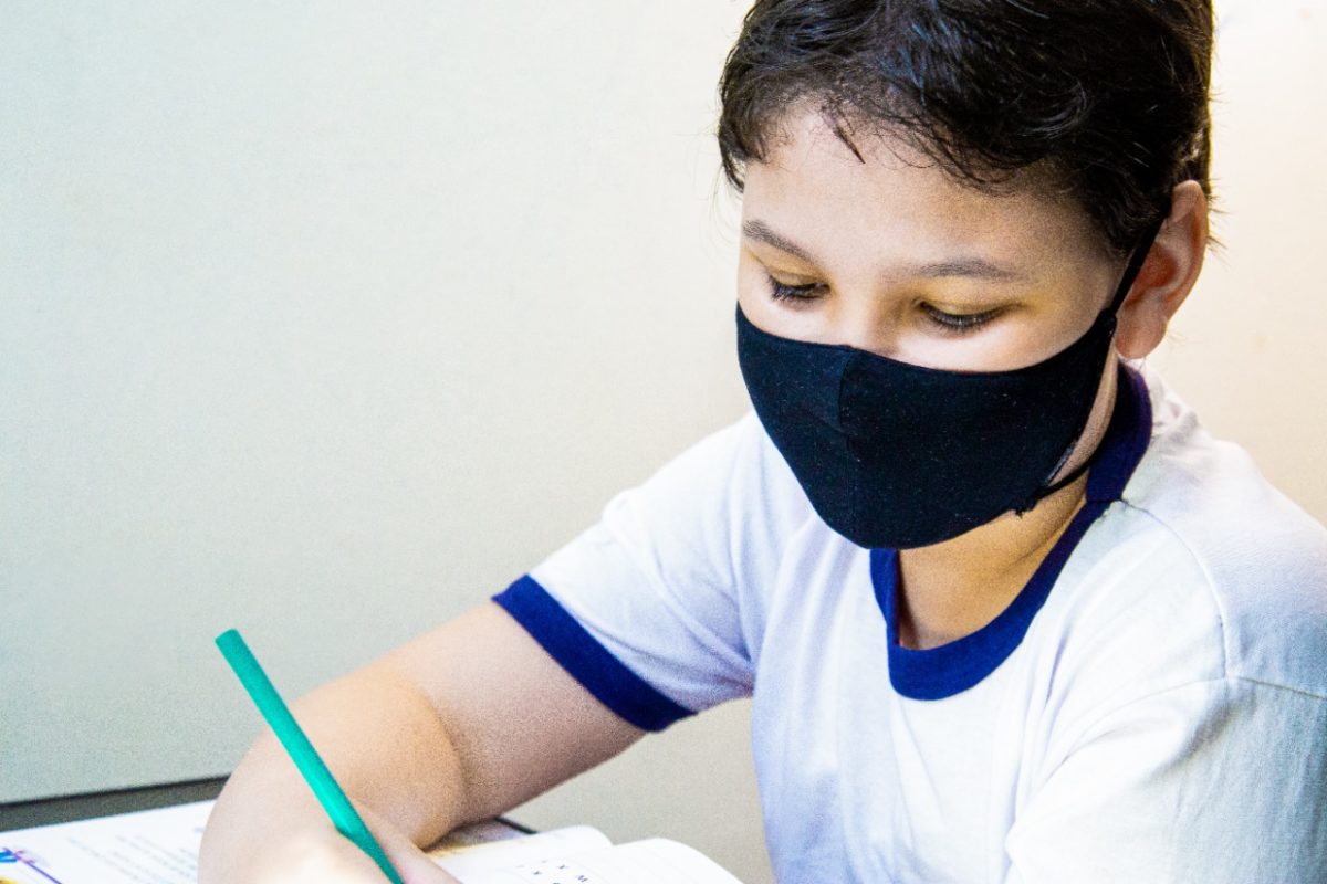 Utilizando máscara preta, um menino branco de camisa branca com bordas na cor azul escuro escreve com lápis verde em um livro.