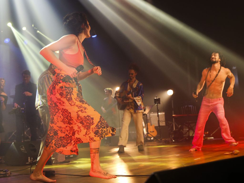 Sobre um palco, numa apresentação de um show, há músicos tocando instrumentos e dois vocalistas cantando e dançando.