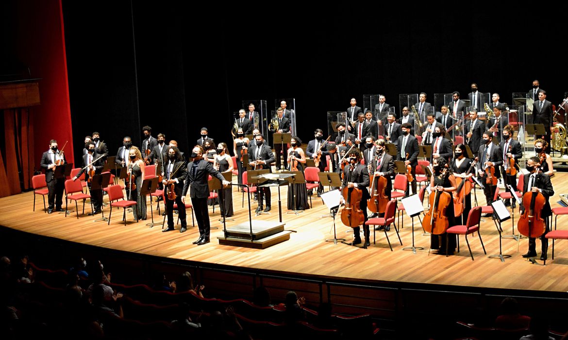 Num palco, integrantes da orquestra estão de pé enquanto seguram seus instrumentos