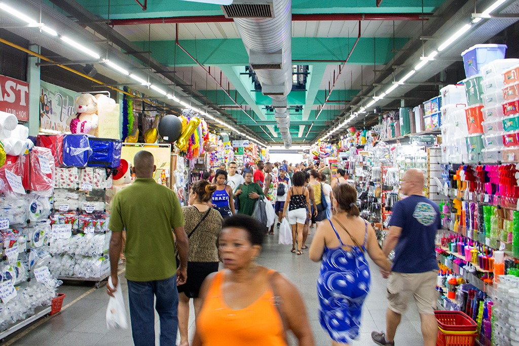 Fotografia do Mercado de Madureira, feita antes da pandemia da COVID-19. Em um ambiente amplo, pessoas caminham por um corredor. Nas bordas do retrato, há prateleiras com produtos variados.