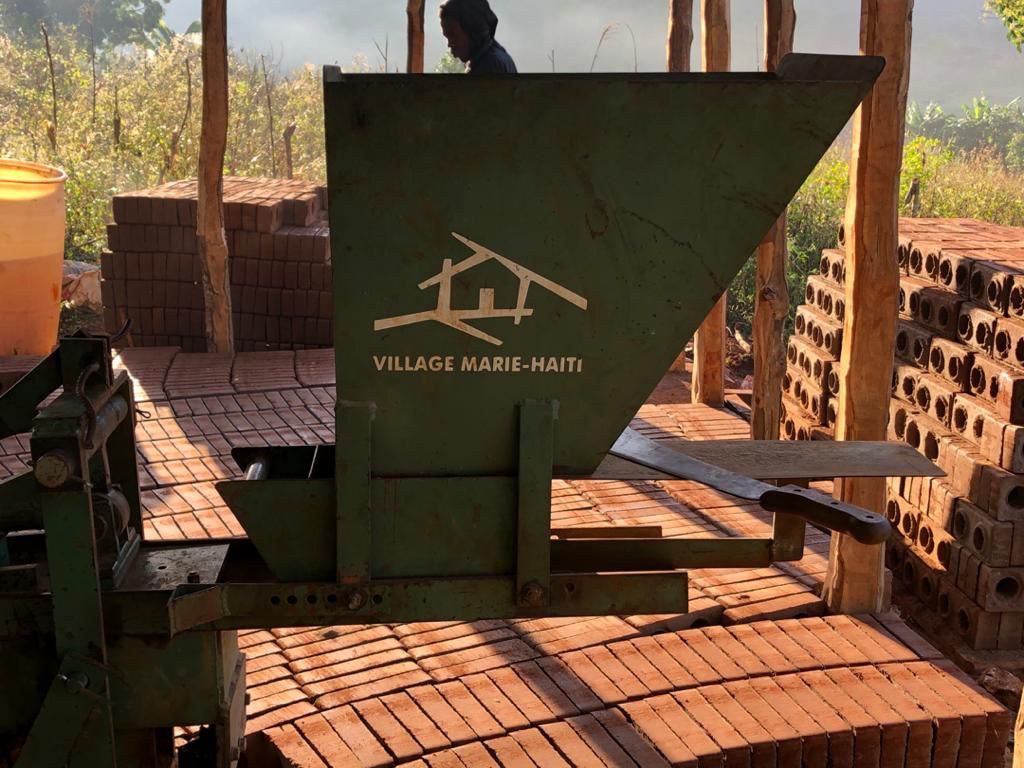 Sobre tijolos ecológicos, há um equipamento de construção civil onde se lê "Village Marie - Haiti"