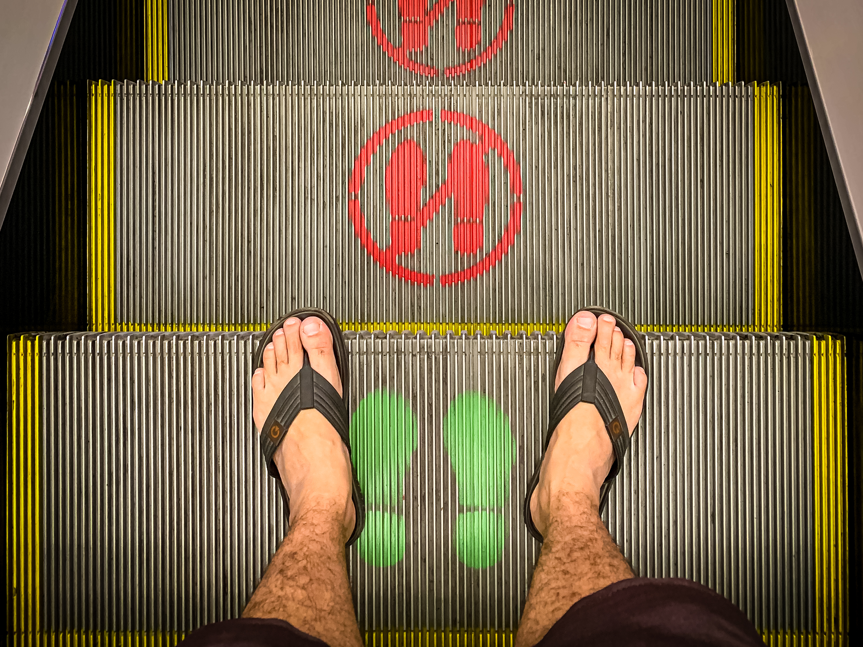 Foto com enfoque nos pés de uma pessoa em uma escada rolante. Na imagem, a pessoa pisa em um degrau com um símbolo verde, os dois degraus seguintes estão marcados com símbolos vermelhos