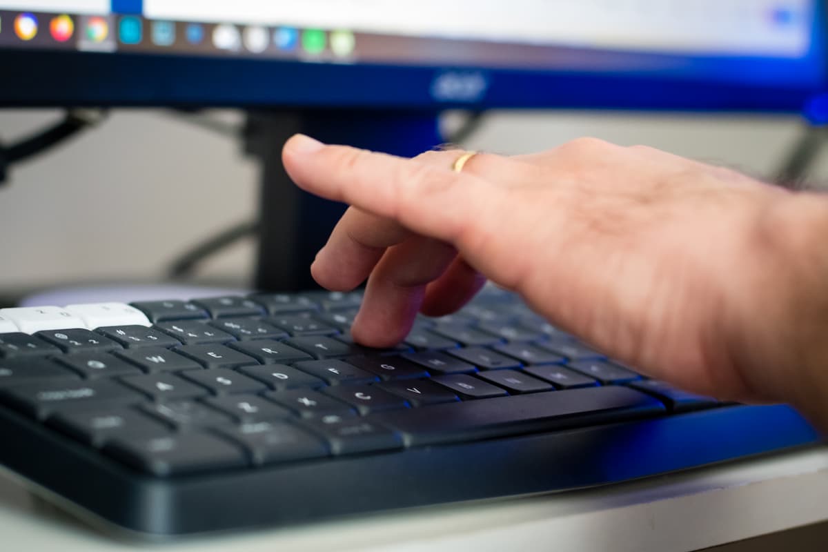 Foto aproximada de um teclado de computador em que uma pessoa digita. Ao fundo, um monitor.