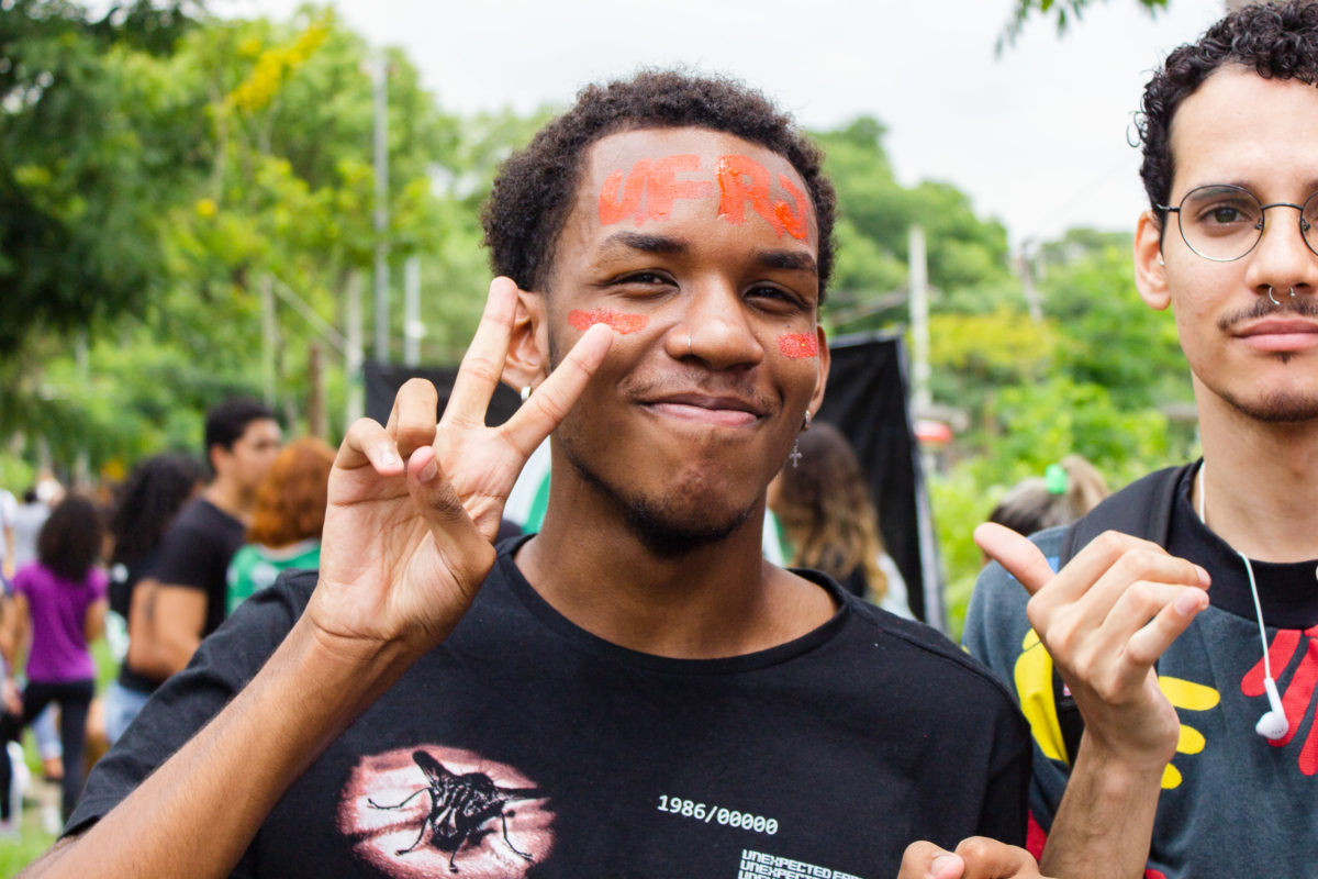 Um jovem negro faz um sinal com os dois dedos das mãos para cima, formando um V. Ele veste uma blusa preta e, em sua testa, está escrito "UFRJ" com tinta laranja. Ao seu lado, um jovem branco, usando óculos.