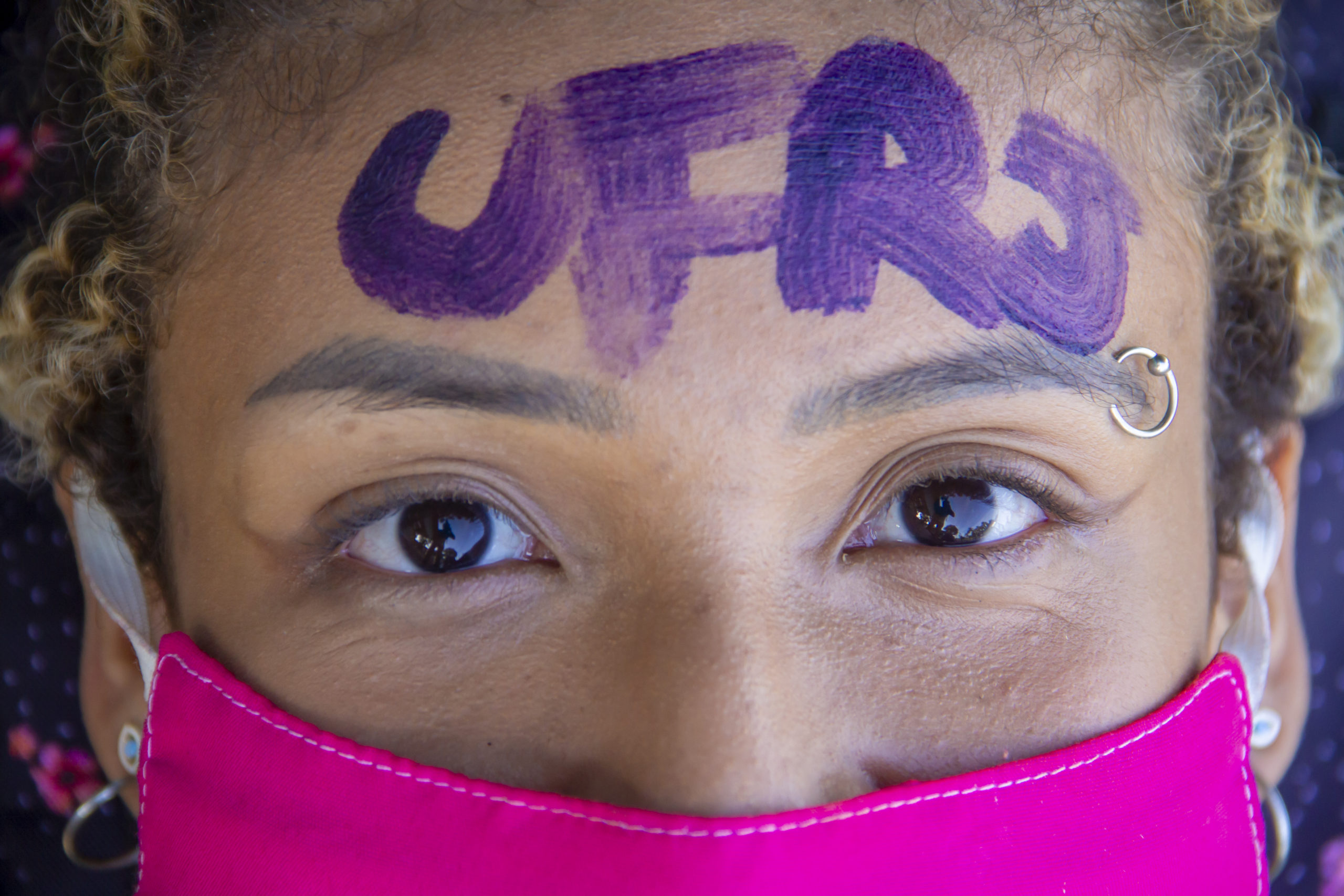 Rosto de uma estudante. Ela usa máscara e tem a palavra UFRJ escrita em sua testa com uma tinta roxa.