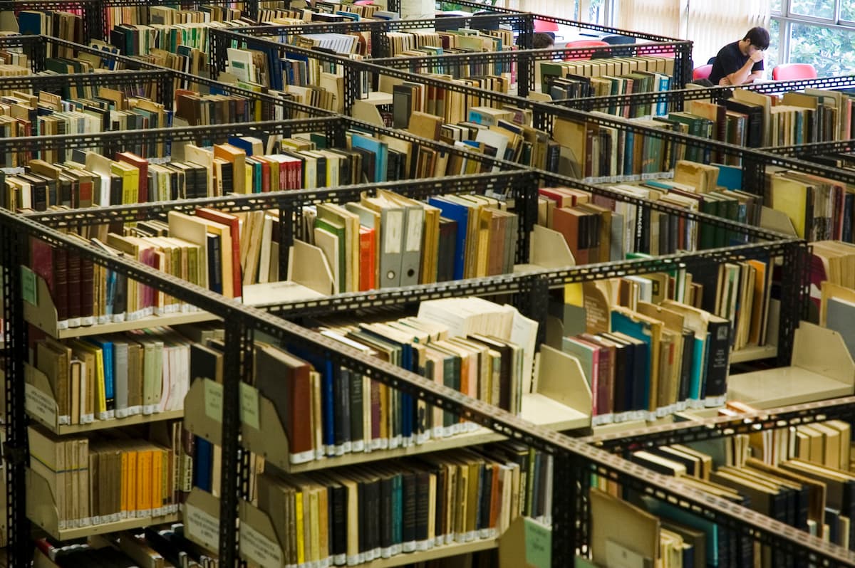 Fotografia mostra fileiras de estantes cheias de livros. No canto direito um aluno estuda.
