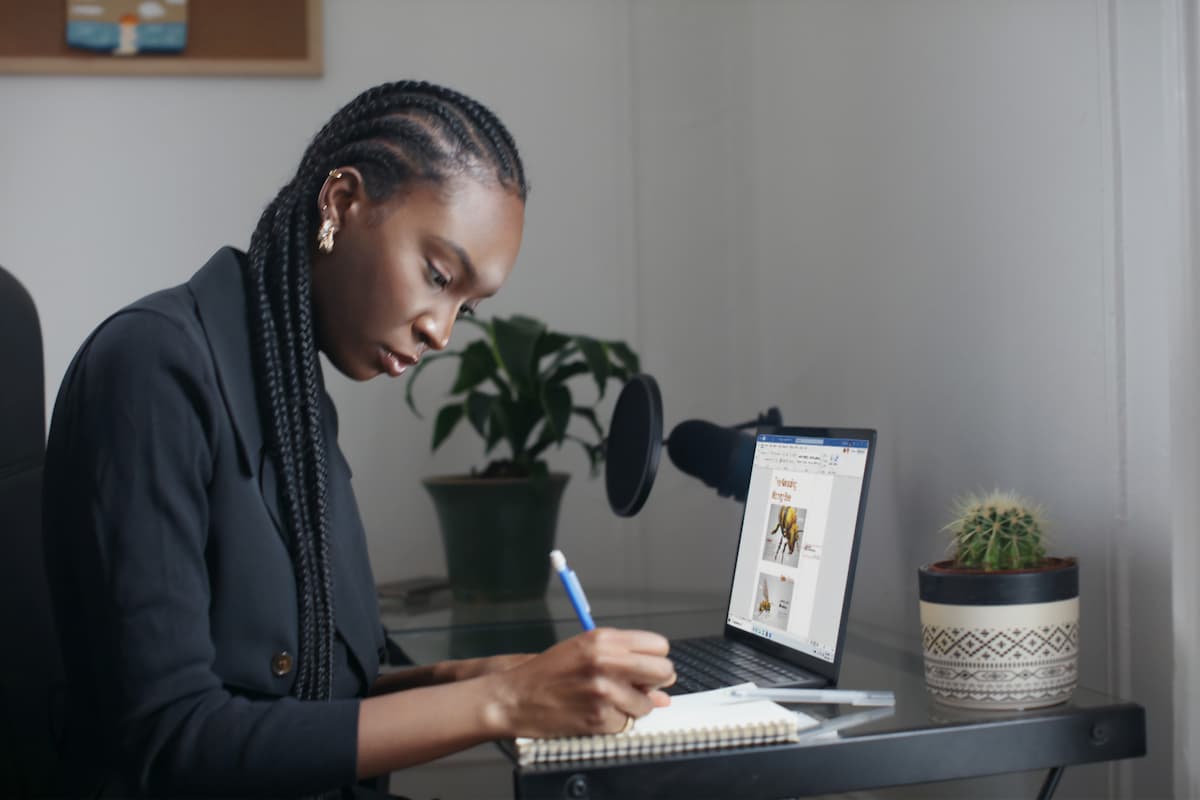 Fotografia de uma mulher negra escrevendo em um caderno. Sobre a mesa ao seu lado, um notebook está aberto.