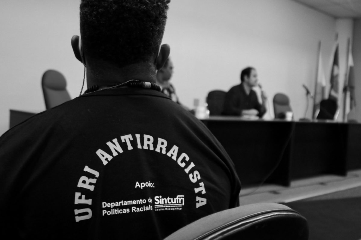 Em primeiro plano, homem assiste a uma palestra. Ele veste uma camisa em que se lê, nas costas, "UFRJ antirracista". Com menor destaque, também está escrito "Apoio: Departamento de Políticas Raciais e Sintufrj".