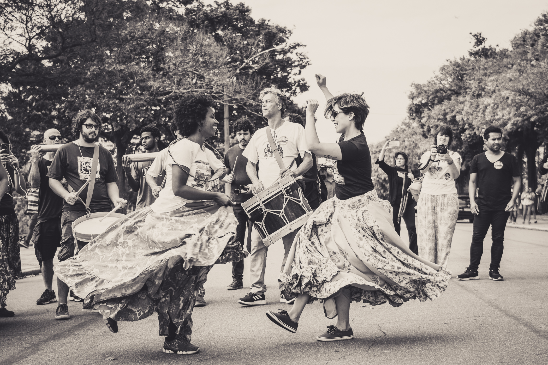duas mulheres de saia comprida dançam na rua, enquanto um grupo toca tambores e fotografa