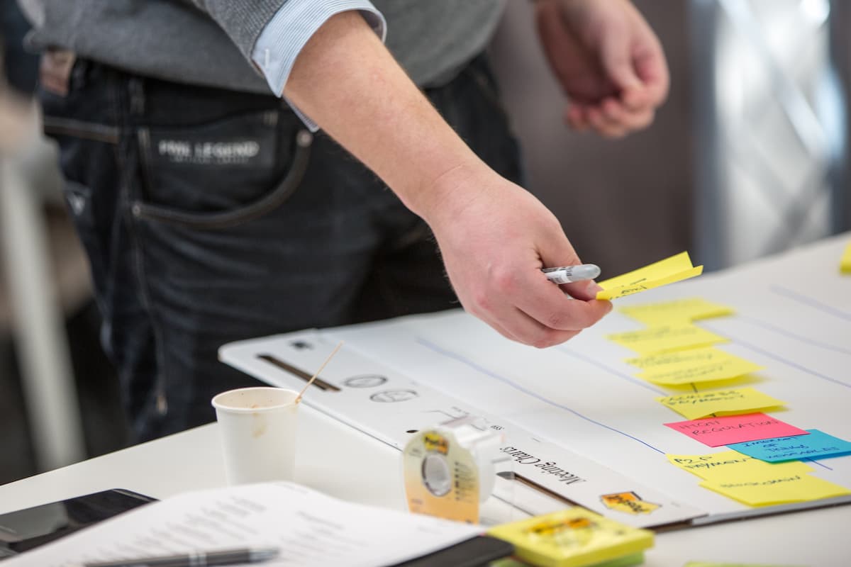 Fotografia mostra as mãos de um homem colando post-its coloridos em um quadro organizacional sobre uma mesa de trabalho