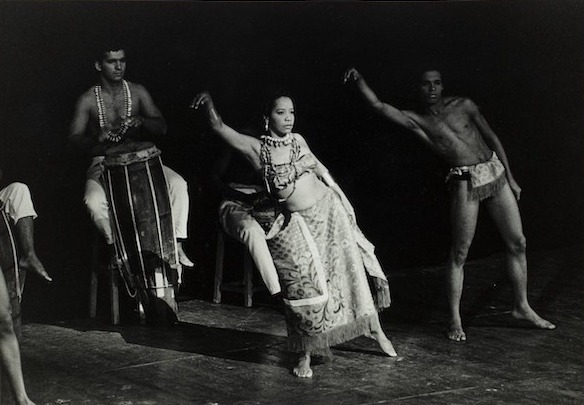 Foto em preto e branco de Mercedes Baptista dançando em um espetáculo de dança afro. Ela usa fios de conta, um top e uma saia longa. Atrás dela, uma pessoa toca atabaque. À sua esquerda, um bailarino a acompanha. 