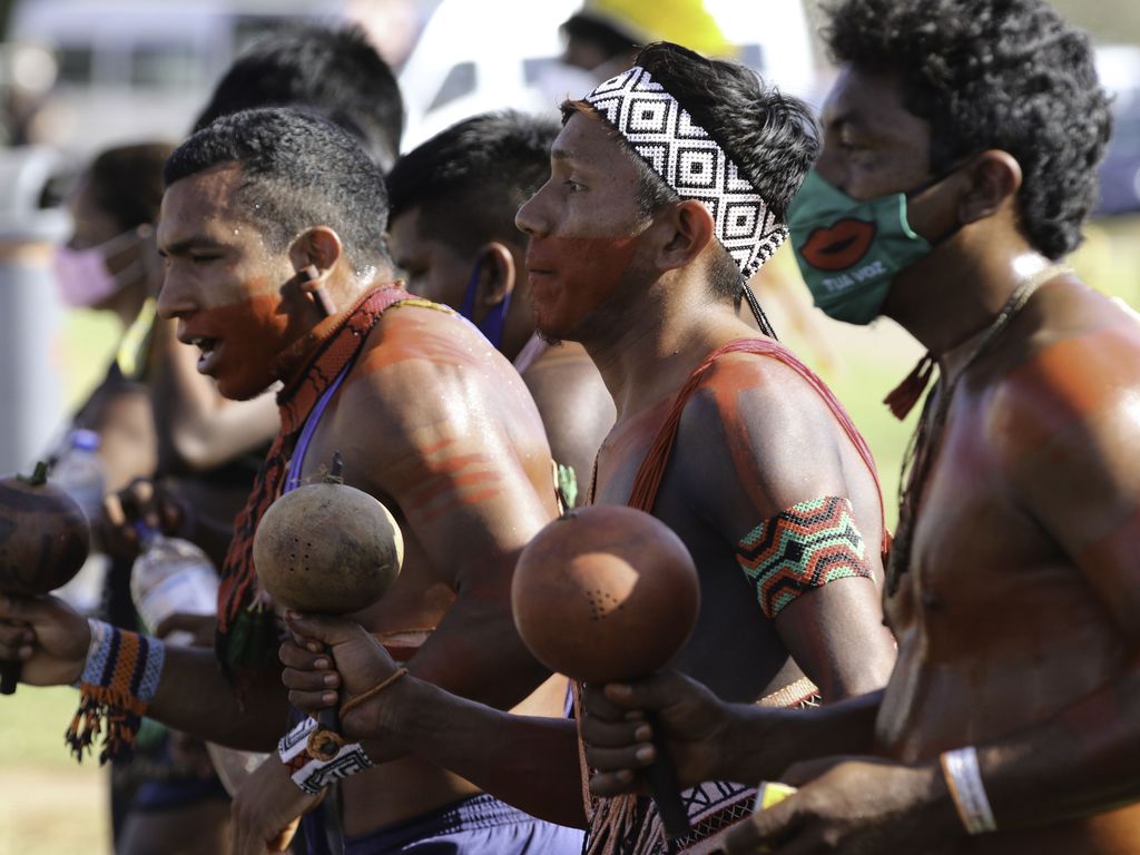 Fotografia de um grupo de indígenas que parece estar dançando. Em destaque estão três deles, homens com pinturas no corpo. Cada um segura um chocalho.