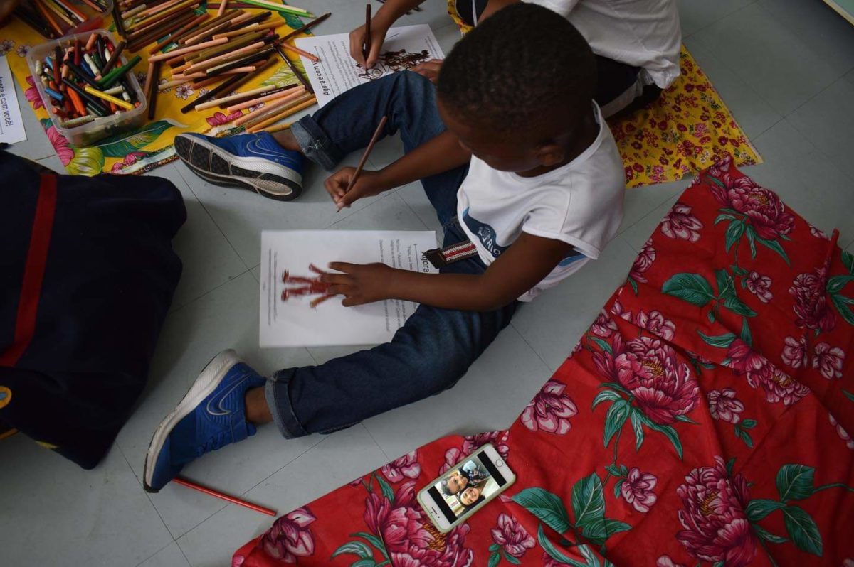 Na foto, um aluno negro está sentado em frente a um desenho segurando um lápis. Em sua volta, podemos ver outra criança e vários lápis de cor espalhados pelo chão e dentro de uma caixa.
