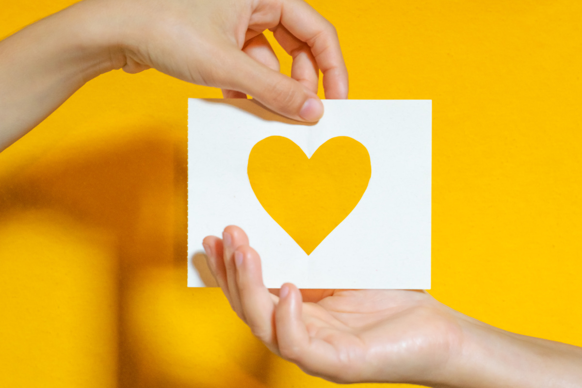 Na foto, duas mãos seguram um papel amarelo cortado em forma de um coração. O fundo da foto é amarelo.