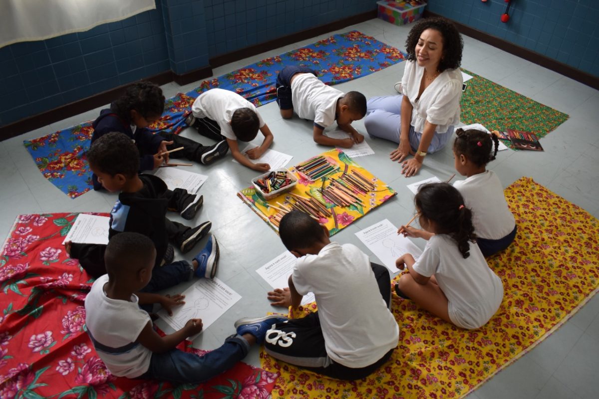 Em uma roda, crianças da rede pública lêem livros infantis. Há uma professora negra com elas.